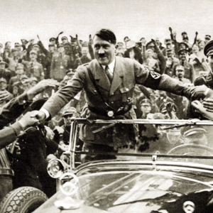 Central figure in a suit with outstretched arms leading a crowd at a historic rally; Nazi swastika armband visible on his sleeve amid cheering supporters.